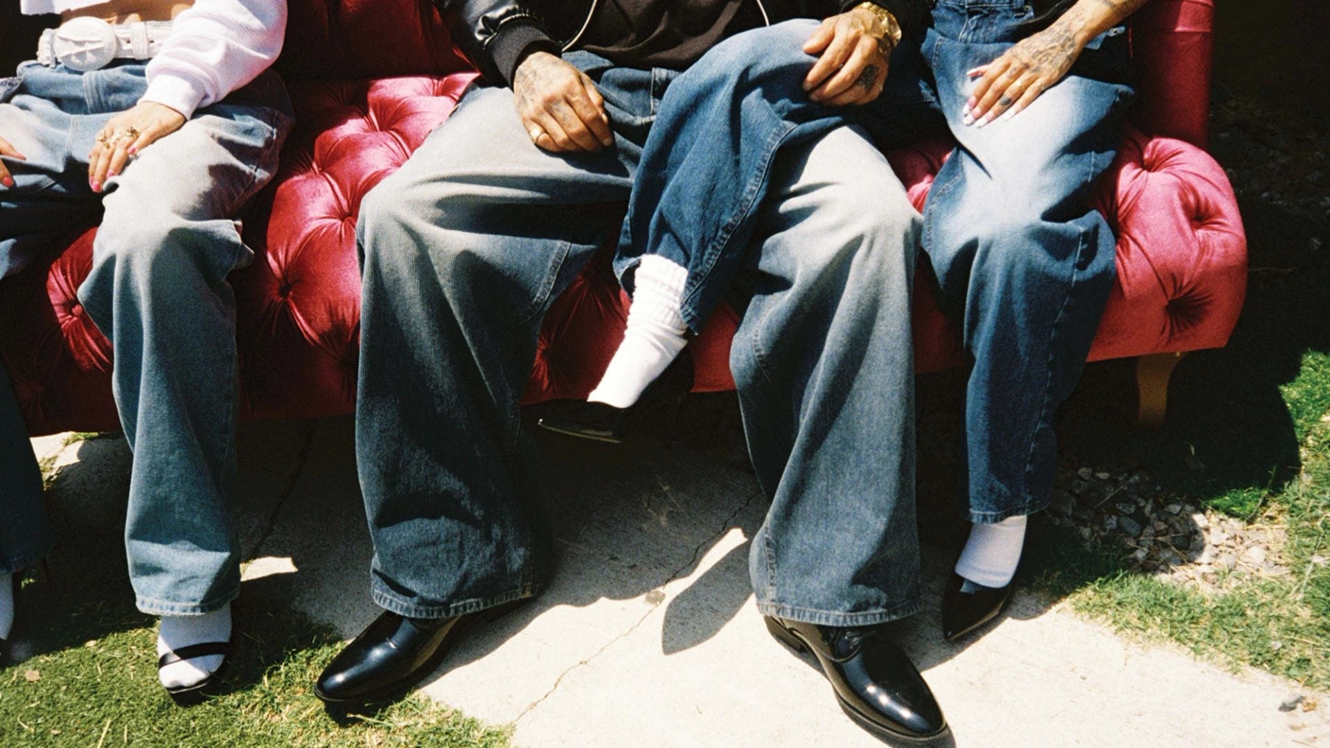 Three people in baggy denim seated on a red velvet couch outdoors