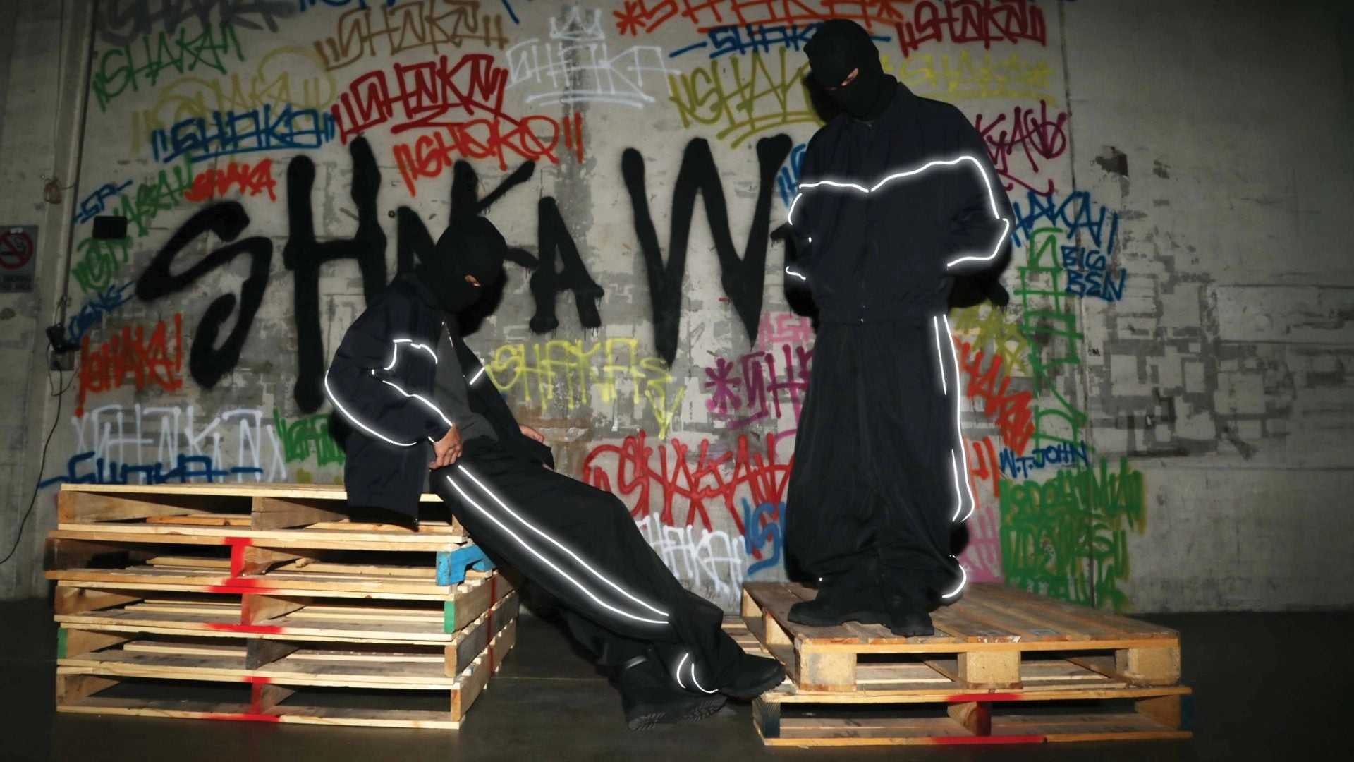 Two models in black tracksuits with reflective piping posing on wood pallets in front of a graffiti wall