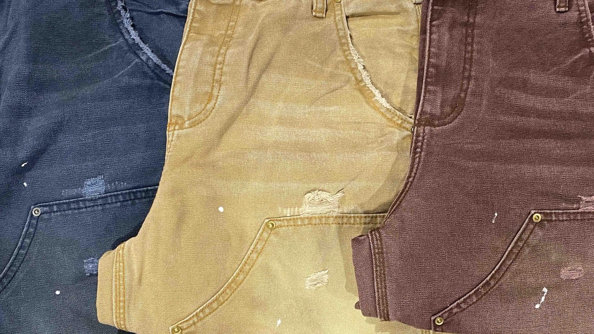 Three colorways of garment-dyed work pants laid side by side: washed navy, tan, and brown